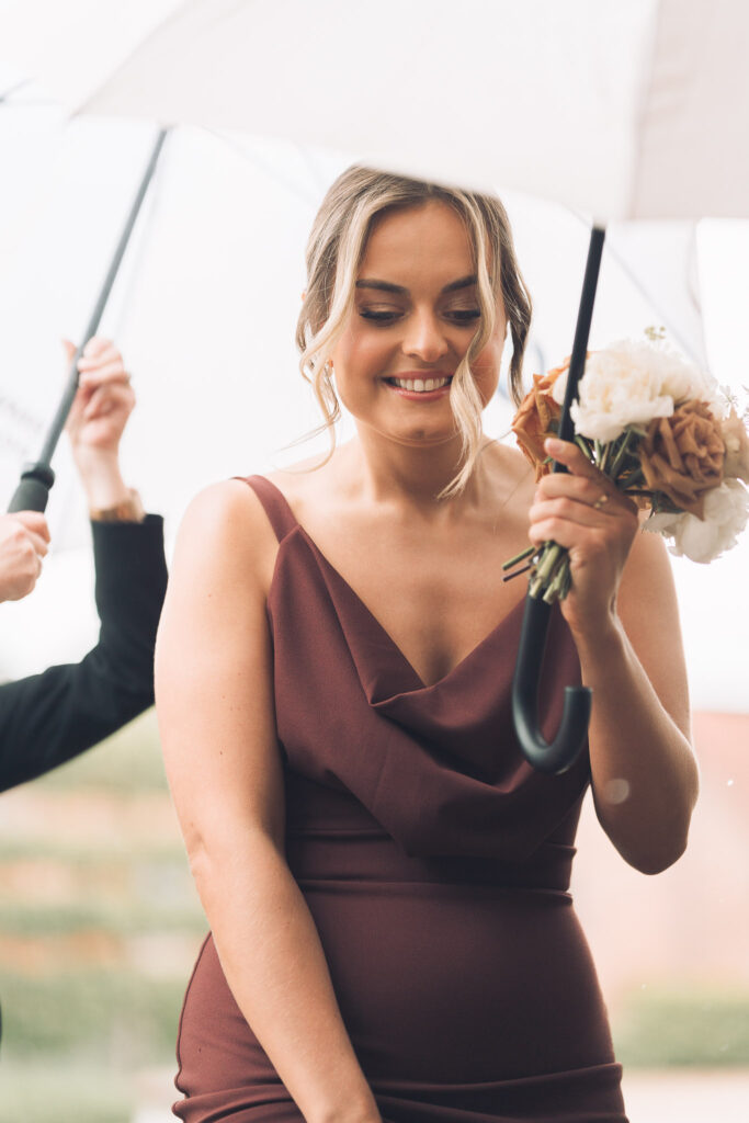 Bridesmaid in elegant cocoa coloured gown carrying small posy of cream and caramel coloured roses and holding a cream umbrella