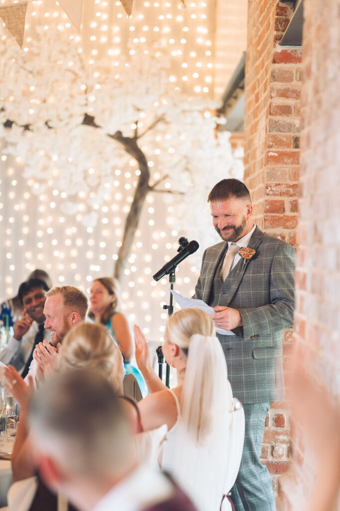 Father of the bride delivering wedding speech surrounded by wedding guests applauding