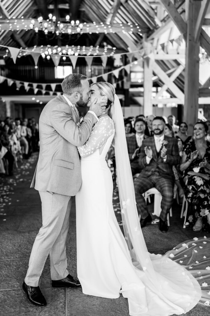 Congregation clapping as bride and groom share first kiss in beamed wedding barn