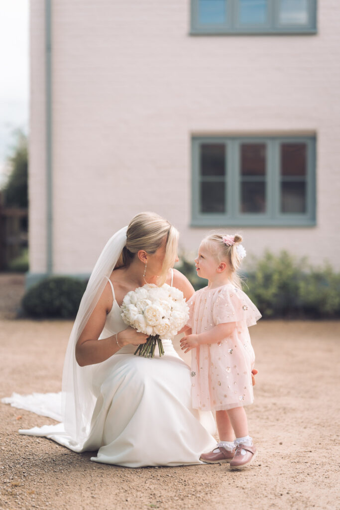 Bride holding large bouquet of cream roses crouching down to greet young guest outside of Rackleys wedding venue