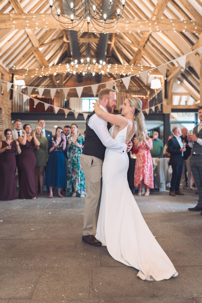 Bride and groom embracing whilst sharing their first dance in front of cheering guests in large beamed wedding barn