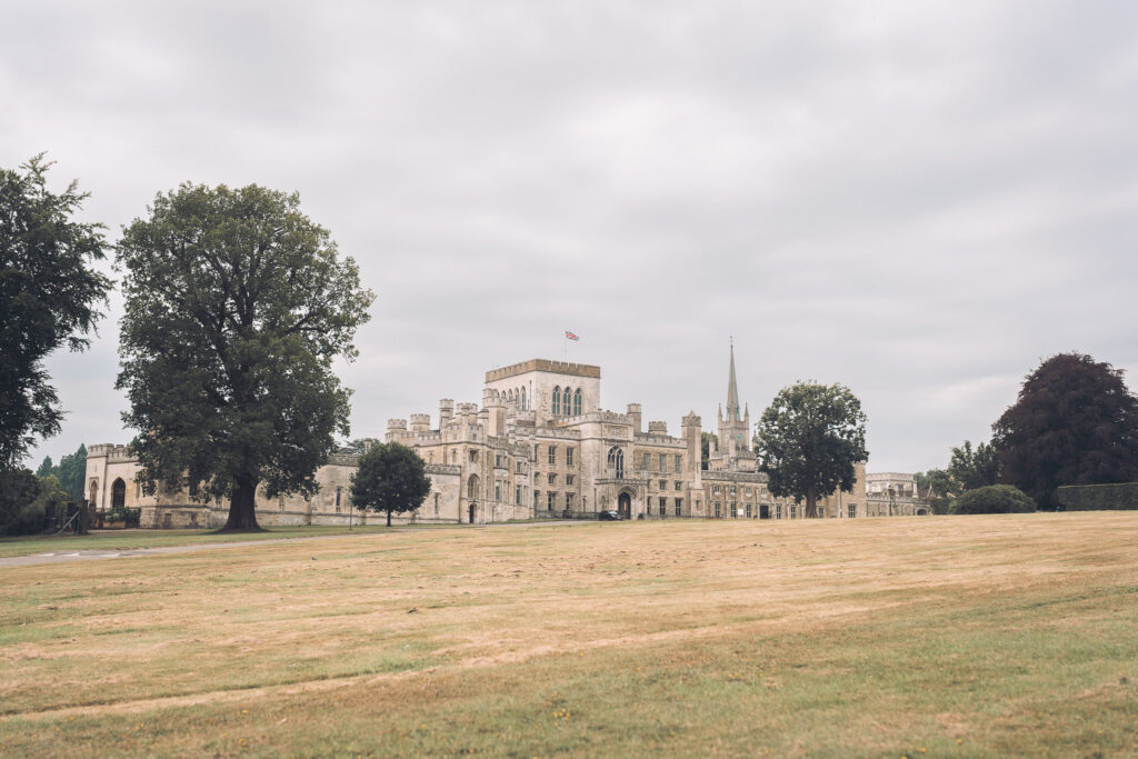 A wide view of a grand, historic stately home with battlements and a prominent spire, set back from a large, dry grassy lawn under a cloudy sky. A Union Jack flag flies from the main building.