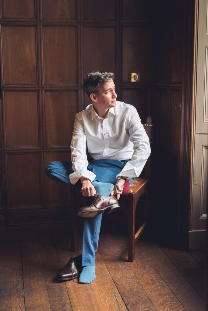 A man wearing a white shirt and blue trousers sitting on a wooden chair, putting on a brown leather shoe in a room with dark wood panelled walls. The man is gazing out through a large window.