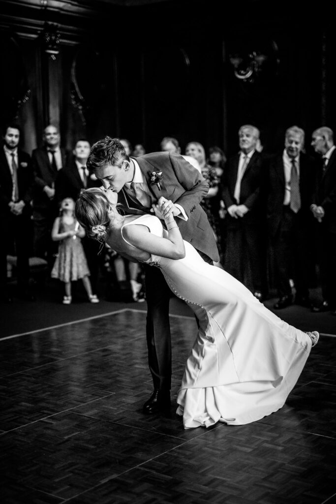 A black and white photo of a couple performing their first dance on a dance floor, with the groom dipping the bride and kissing her as guests watch in the background.