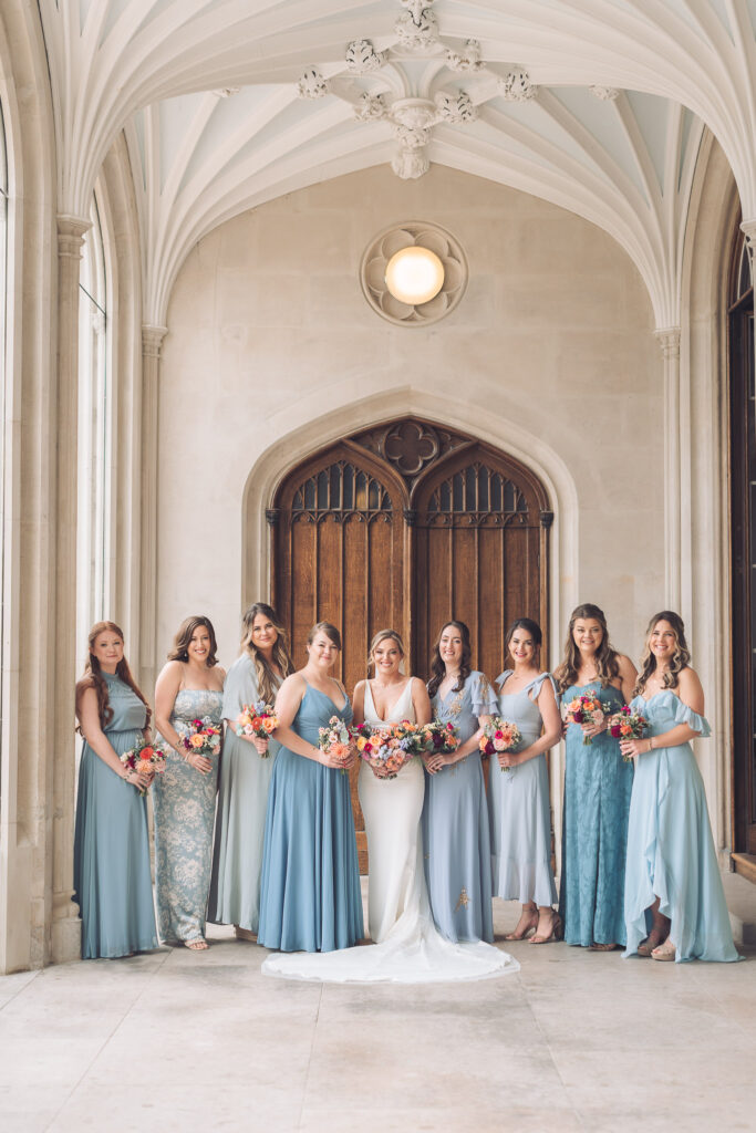 A bride in a white gown and her eight bridesmaids, all wearing long dresses in shades of blue, standing in a line under a stone archway with gothic-style architecture and holding bright bouquets.