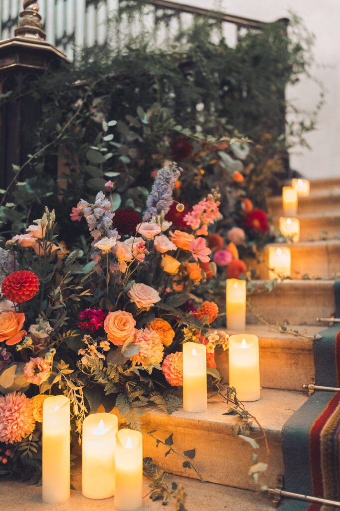 A close-up of a staircase decorated with lush floral arrangements featuring pink, coral, and red flowers, and illuminated by rows of tall, flickering LED candles.