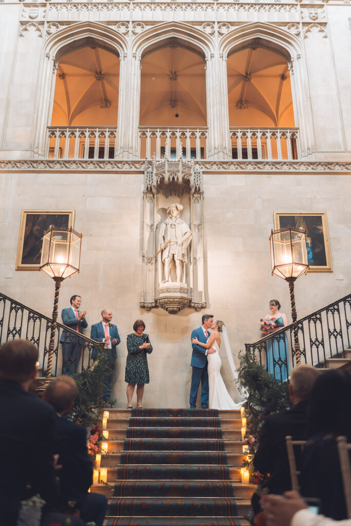 A couple kissing during a wedding ceremony on a grand, carpeted staircase inside a stately hall. Guests stand on the stairs and watch, and a statue is set into the wall above.