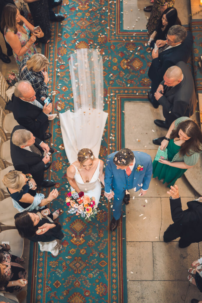 An aerial view looking down at a bride in a white gown and a groom in a blue suit walking down a turquoise patterned runner on a staircase as confetti falls, surrounded by applauding guests.