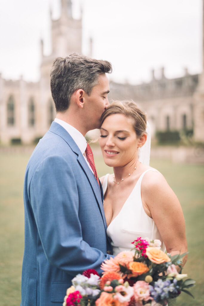 A close-up of a couple embracing outdoors on a lawn with a stone stately home in the blurred background. The groom, in a blue suit, is kissing the bride on the forehead. She is holding a large, colorful bouquet.