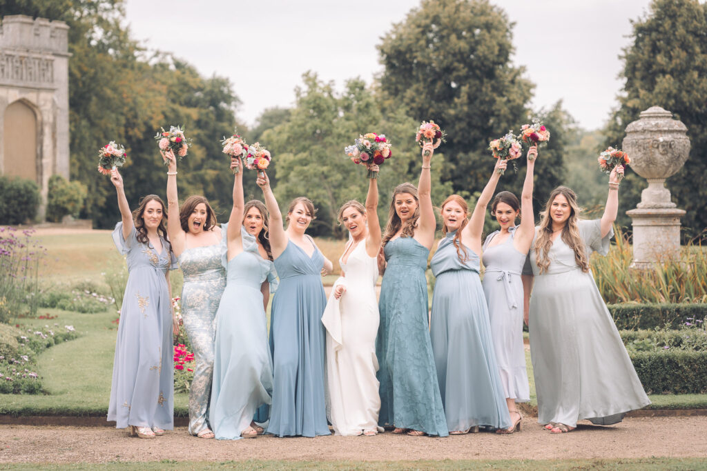 A bride and eight bridesmaids, all holding bouquets high in the air, standing on a gravel path in a garden setting with trees and the stately home in the background.