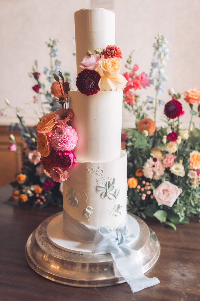 A three-tier white wedding cake decorated with fresh dahlias and roses in shades of pink, orange, and maroon, and light blue piping details on the bottom tier. A pale blue ribbon is tied around the base.