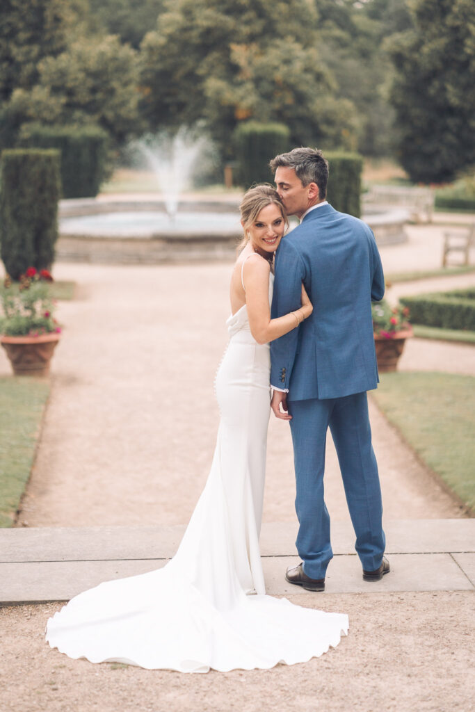 A full-length portrait of a couple outdoors in a manicured garden. The groom, in a blue suit, is kissing the bride on the forehead as she looks back over her shoulder and smiles. A fountain is visible in the background.